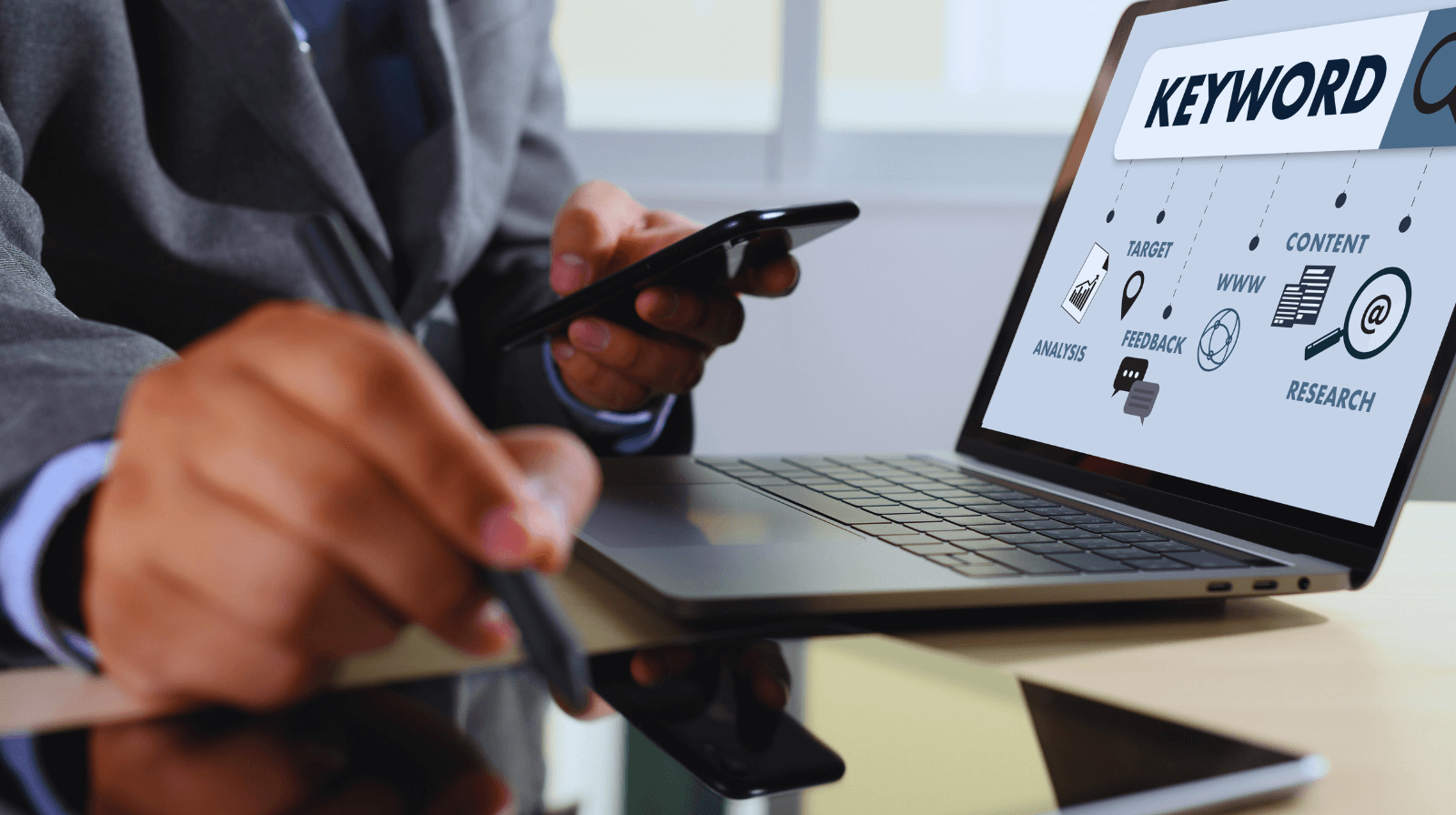 Man using smartphone beside laptop showing ‘Keyword’ and icons, illustrating the importance of keywords for SEO